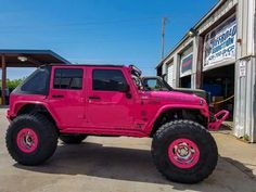 a bright pink jeep parked in front of a building with large tires on it's wheels