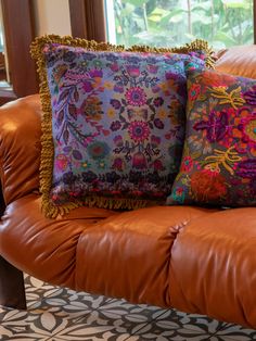 two colorful pillows sitting on top of a brown leather couch in front of a window