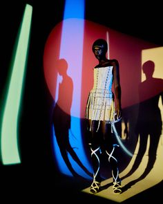 a woman is standing in front of a colorful wall with shadows on it and wearing a white dress