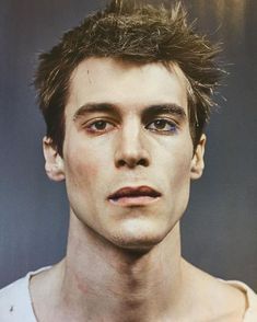 a man with short hair and blue eyes looks at the camera while wearing a white tank top