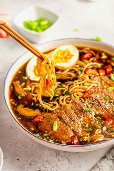 someone holding chopsticks over a bowl of beef ramen with an egg on top