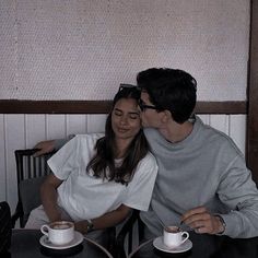 a man and woman sitting next to each other at a table with two cups of coffee