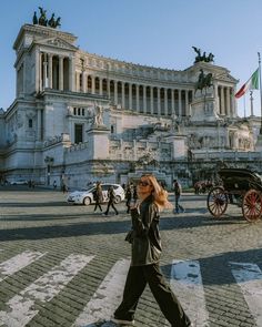 a woman walking across a cross walk in front of a large building with statues on it