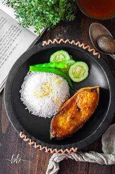 fish, rice and cucumbers on a black plate next to an open book