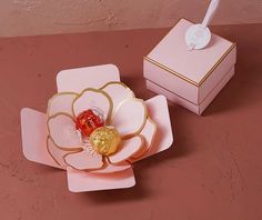 a pink box with a gold flower on top and a candy in the middle sitting next to it