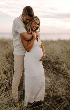 a pregnant woman standing next to a man on top of a grass covered field in front of the ocean