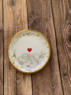 a white and gold plate with a red heart painted on the side sitting on a wooden surface