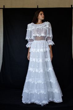 a woman standing in front of a black backdrop wearing a white dress with sheer sleeves