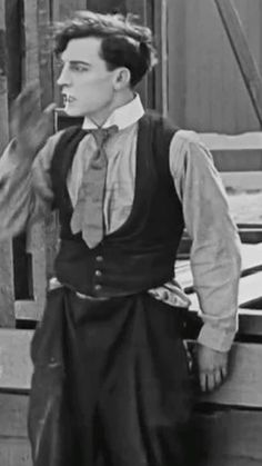 an old photo of a man wearing a vest and tie standing in front of a fence