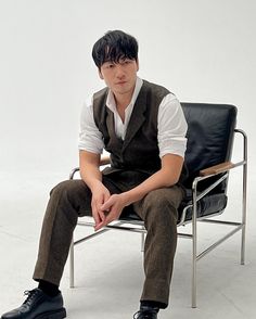 a young man sitting on top of a black chair next to a white wall with his hands in his pockets