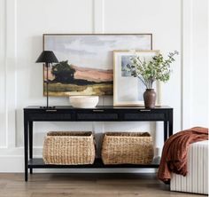 a black console table with two baskets underneath it and a painting on the wall behind it