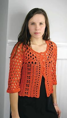 a young woman wearing an orange crochet jacket and black dress standing in front of a white door