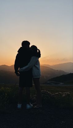 two people standing on top of a hill with their arms around each other as the sun sets