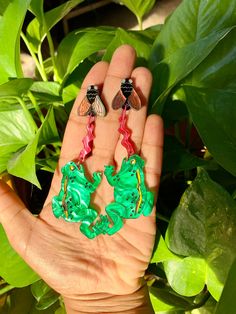 a hand holding two green and pink earrings in it's palm, next to some leaves