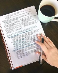 a woman's hand is holding a pen and writing on a book next to a cup of coffee