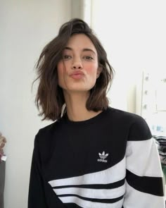 a woman with short brown hair wearing a black and white adidas sweater in front of a window