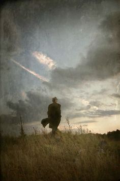 a man standing on top of a grass covered hill under a cloudy sky with clouds