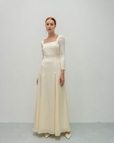 a woman standing in front of a white wall wearing a long cream dress and earrings