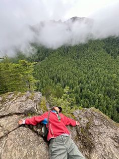 a man laying on top of a large rock next to a lush green forest covered hillside