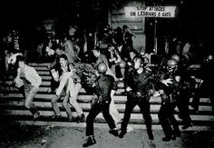 black and white photograph of police running down stairs in front of people with riot gear