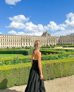 a woman standing in front of a large building with hedges and bushes around her, wearing a long black dress