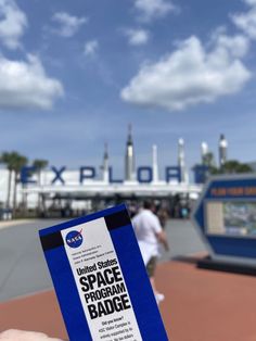 someone holding up a space program badge in front of a building with people walking around