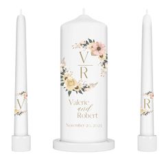 three white candles with floral designs on the top and bottom, one candle has a gold initials