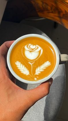 a hand holding a white cup filled with orange liquid and topped with a rose design