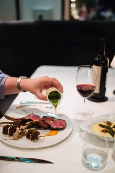 a person pouring sauce on a plate with food in front of them and a glass of wine to the side