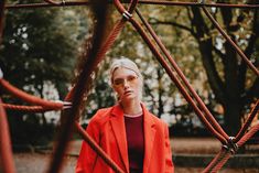 a woman in an orange jacket and sunglasses standing next to a rope structure with trees behind her