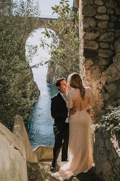 a bride and groom are standing on the stairs by the water in front of an old bridge