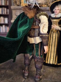 a doll is dressed in medieval clothing and holding a green cape while standing next to a bookshelf