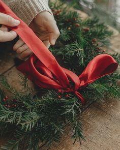 a person tying a red ribbon around a wreath on a wooden table with greenery