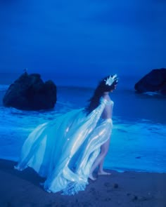 a woman in white dress standing on beach next to ocean with rocks and blue sky