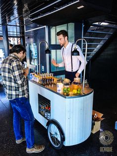 two men standing in front of a food cart with drinks on the counter and bottles behind it