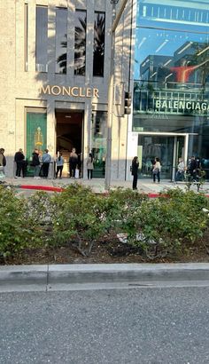 people are standing in front of a building with many windows and plants on the sidewalk