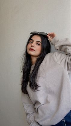 a woman with long black hair wearing a gray sweatshirt and jeans, leaning against a wall