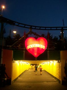 the entrance to an amusement park at night with people walking under it and a giant heart sign