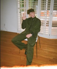 a young man sitting on a chair with his hand up in the air while wearing an army green suit