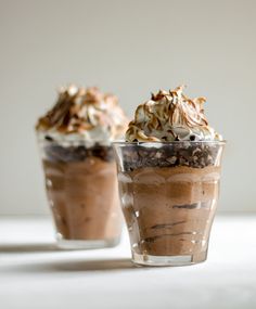 two dessert cups with whipped cream and chocolate toppings on top, sitting on a white surface