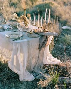 a table set with candles and plates in the middle of some tall grass, surrounded by dry brush