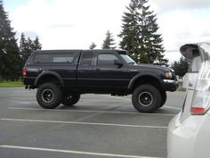 a large black truck parked in a parking lot next to a white car and trees