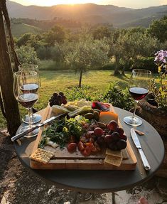 a table with wine glasses, cheese and fruit on it in front of an outdoor setting