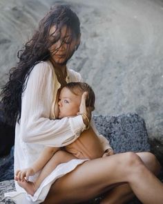 a woman holding a baby sitting on top of a rocky beach next to the ocean