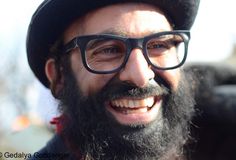 a close up of a person wearing glasses and a hat with a beard smiling at the camera