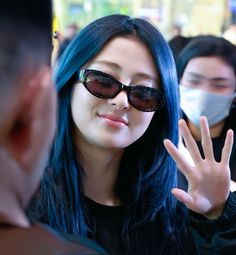 a woman with blue hair wearing sunglasses and a face mask is waving at someone behind her