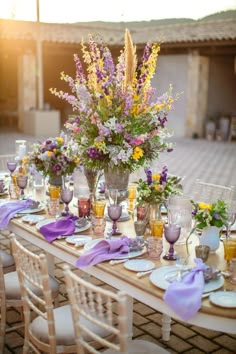 a table set up with purple and yellow flowers in vases, plates and napkins