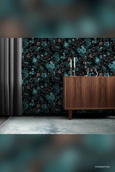 A luxurious interior featuring a dark floral wallpaper with mint green and teal flowers against a black background. The room includes a wooden sideboard topped with modern candle holders and a decorative black bull figurine. Grey curtains and a light, textured rug complete the elegant look Black Floral Wallpaper, Wallpaper In Black, Mint Green Wallpaper, Feature Walls, Textile Artist