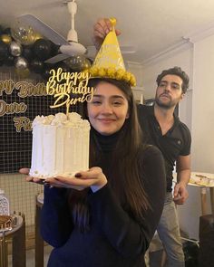 a woman holding a birthday cake in front of a man with a party hat on