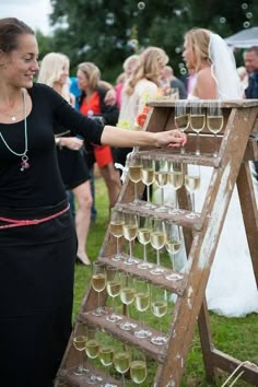 a woman standing next to a wooden ladder filled with wine glasses on top of a grass covered field